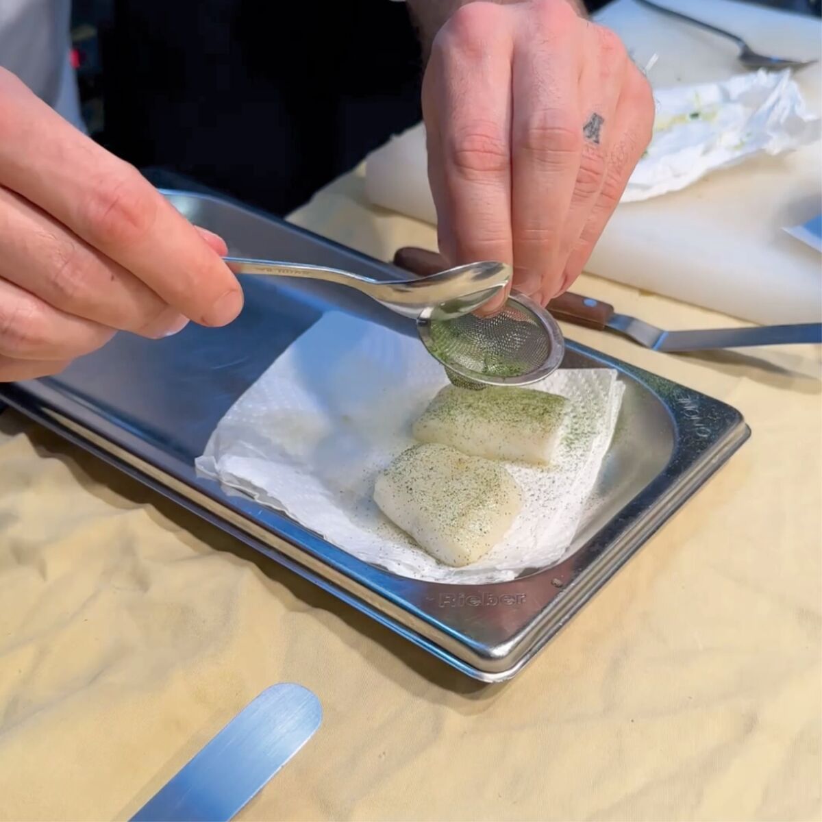 Koch streut grünes Pulver auf zwei Stück Fischfilet auf Metalltablett